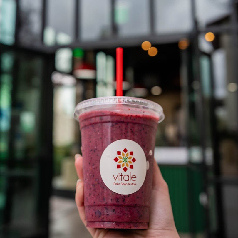 Hand holding a Vitale Poke Shop & More berry smoothie with a red straw, storefront blurred in background.