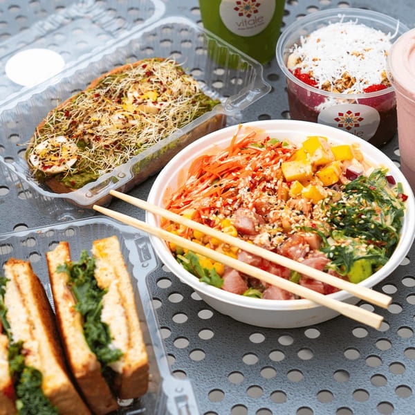 Vitale takeout containers with avocado toast, poke bowl, acai bowl, and green juice on a metal table.