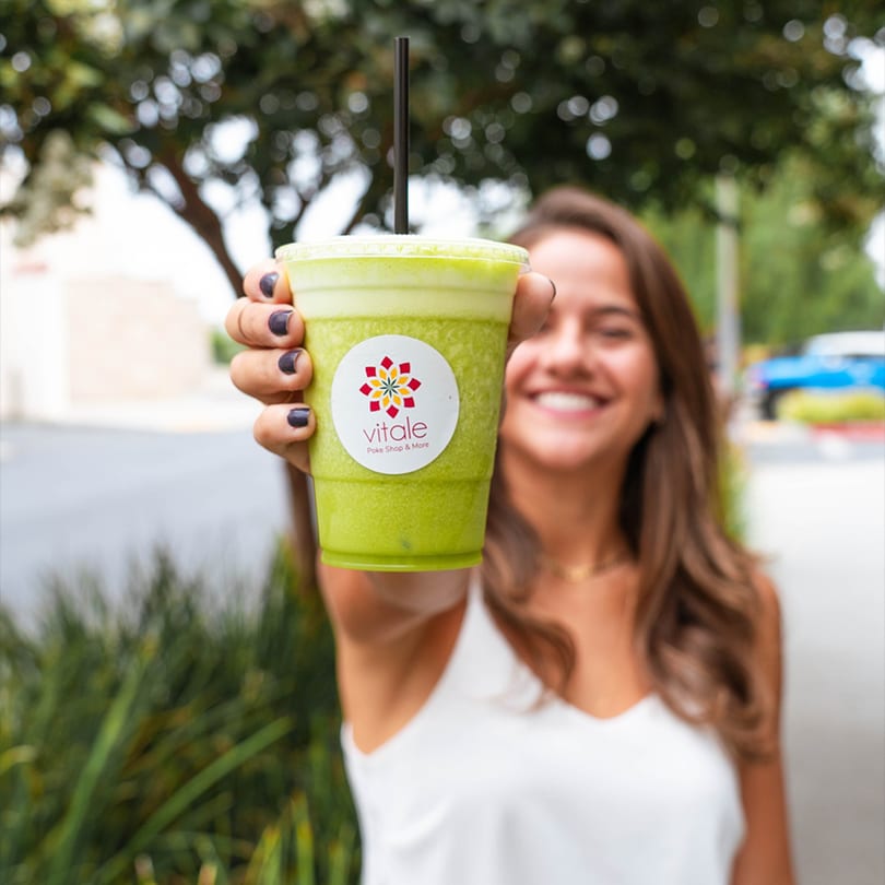 Smiling woman holding up a green smoothie with a Vitale Poke Shop & More logo.