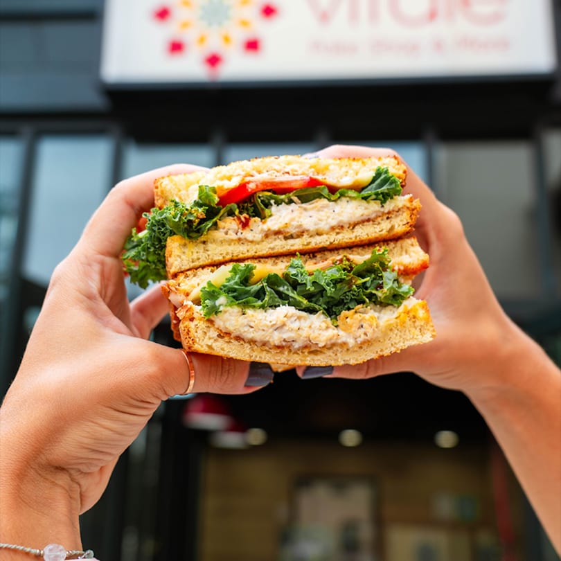 Hands holding a grilled sandwich filled with chicken salad, kale, and tomato, in front of a restaurant sign.