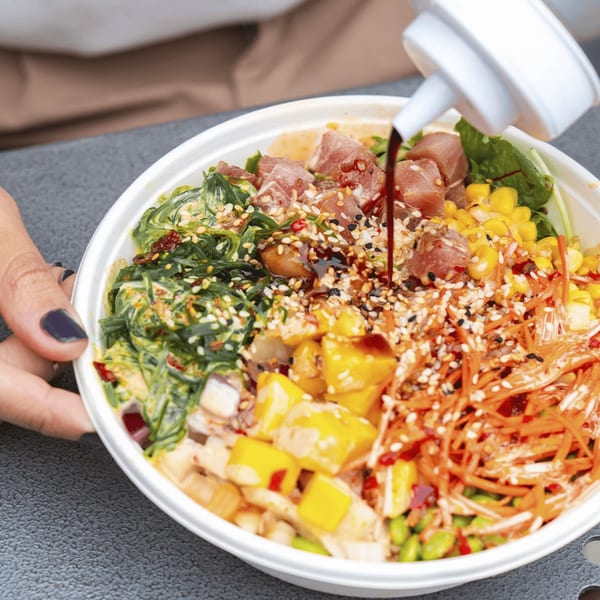 A person's hand holds a poke bowl as sauce is drizzled over the colorful ingredients.