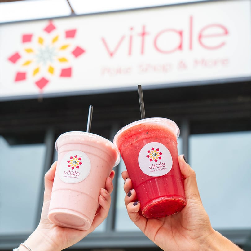Two hands holding up Vitale smoothies, one pink and one red, in front of the restaurant sign.