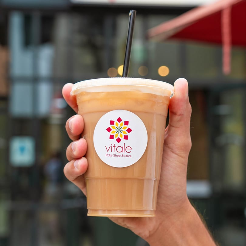 Hand holding a Vitale Poke Shop & More iced drink with a black straw outdoors.