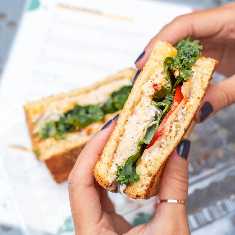 Hands holding a grilled sandwich with chicken salad, kale, and tomato, with another sandwich in the background.