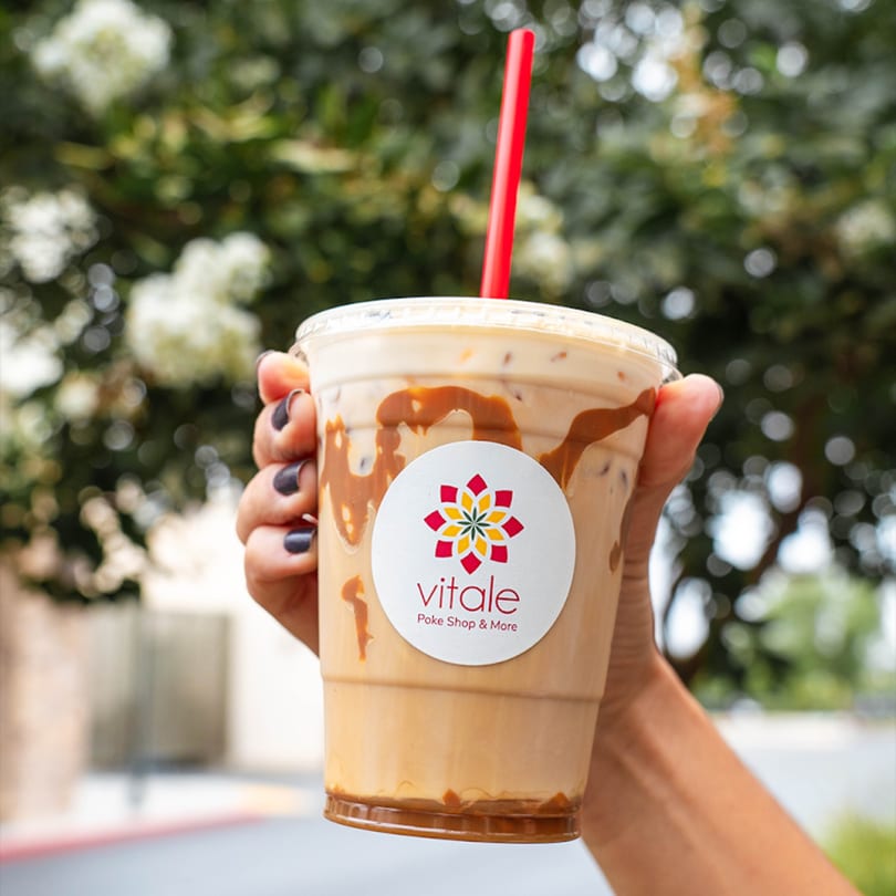 Hand holding a Vitale Poke Shop & More iced coffee with caramel drizzle and a red straw.