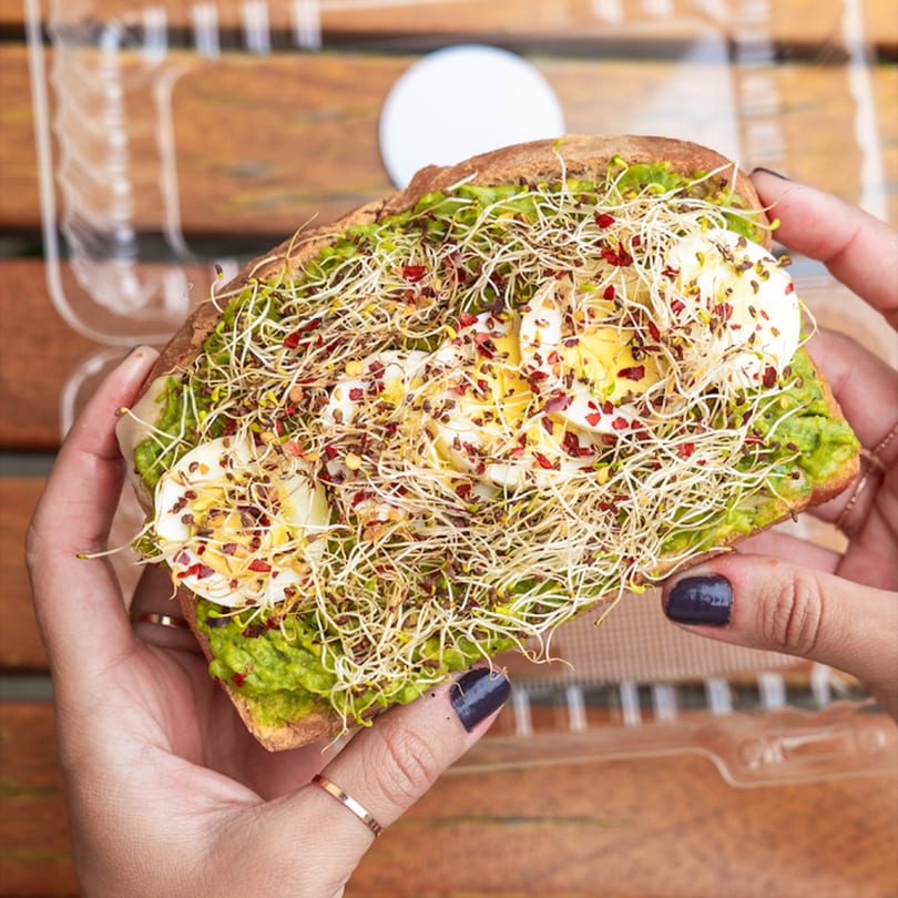 Hands holding avocado toast topped with sliced eggs, sprouts, and red pepper flakes.