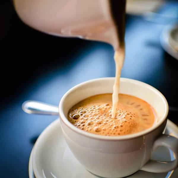 Milk being poured from a pitcher into a white cup of coffee, creating foam.