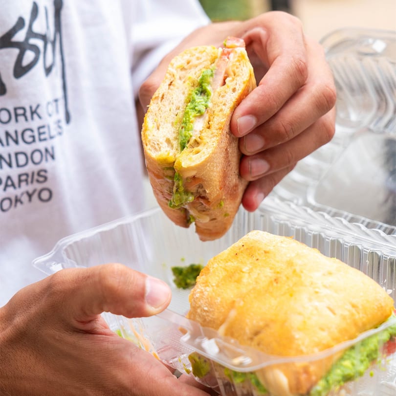 Hands holding a sliced sandwich with green avocado spread, with another sandwich in a plastic container.